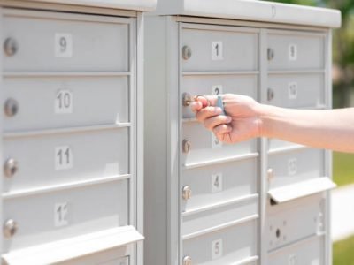 Person's hand holding key unlock-lock mailbox by house number, selective focus. Receiver gets mail and small parcel from the row of outdoor metal storage, depth of field. Man close residential letterbox.